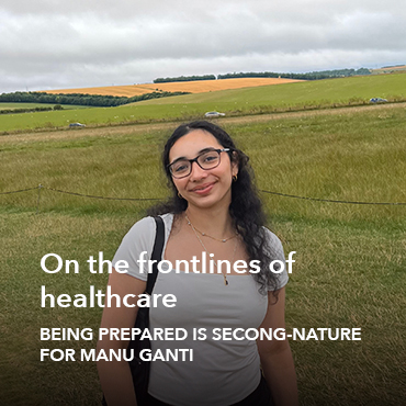 A young woman with long, curly dark hair and glasses, wearing a white top and a backpack by one shoulder strap, standing before a green field with yellow flowers.